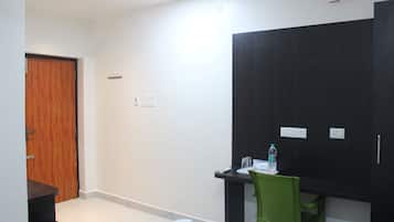 A hotel room with a wooden door, a desk with a chair, a black wardrobe, and a small table with a water bottle.
