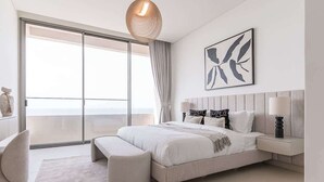 Une chambre moderne avec un grand lit, une table de chevet, un banc, un luminaire suspendu et une grande fenêtre donnant sur un beau paysage.
