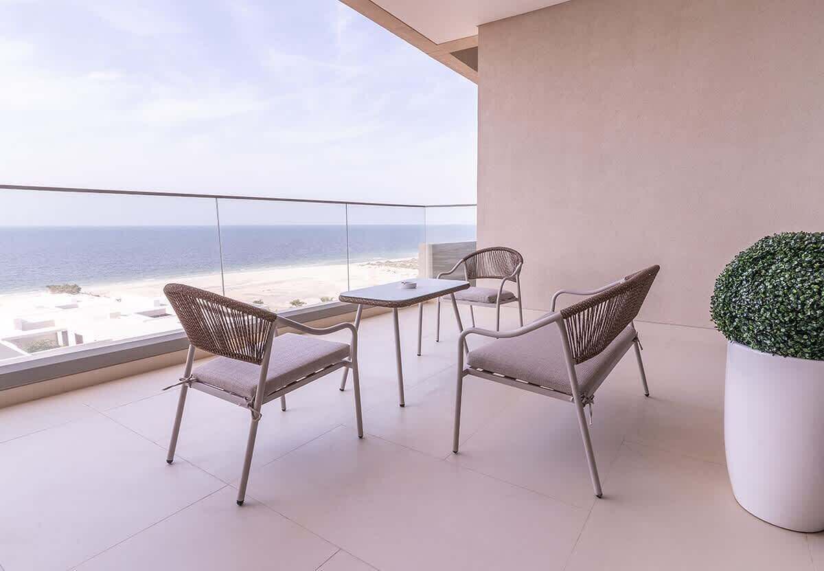 Uma varanda com vista para a praia, duas cadeiras de vime, uma mesinha e um vaso de planta.
