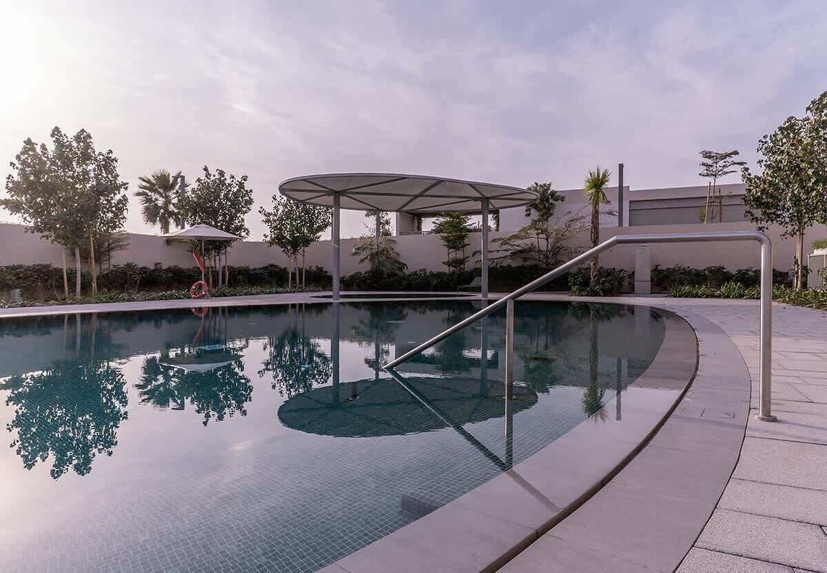 Une piscine à débordement avec une bordure en verre, entourée d’une zone pavée, d’arbres et d’un bâtiment en arrière-plan.
