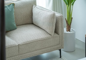 Un sofá color beige con un cojín verde, una planta en maceta y un objeto cilíndrico blanco.