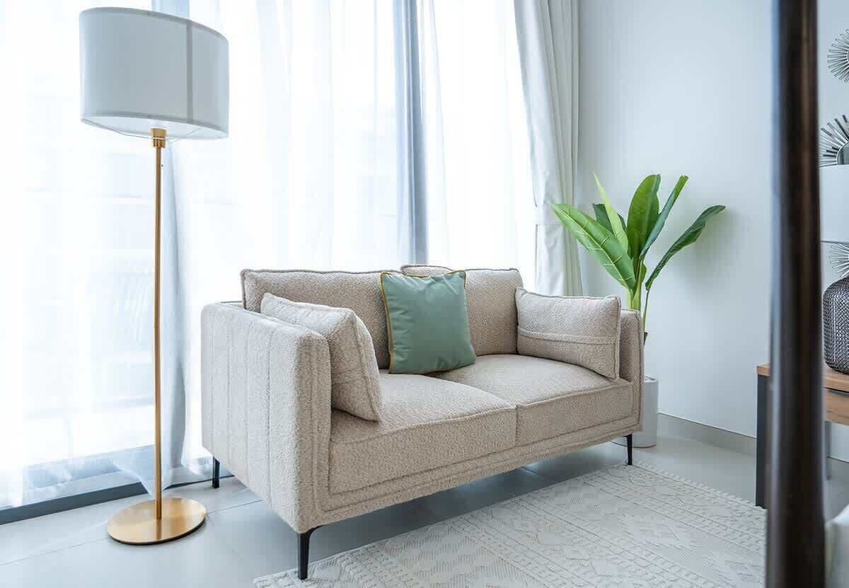 A beige sofa with a gold floor lamp, a green pillow, and a plant in a white room with a patterned rug.