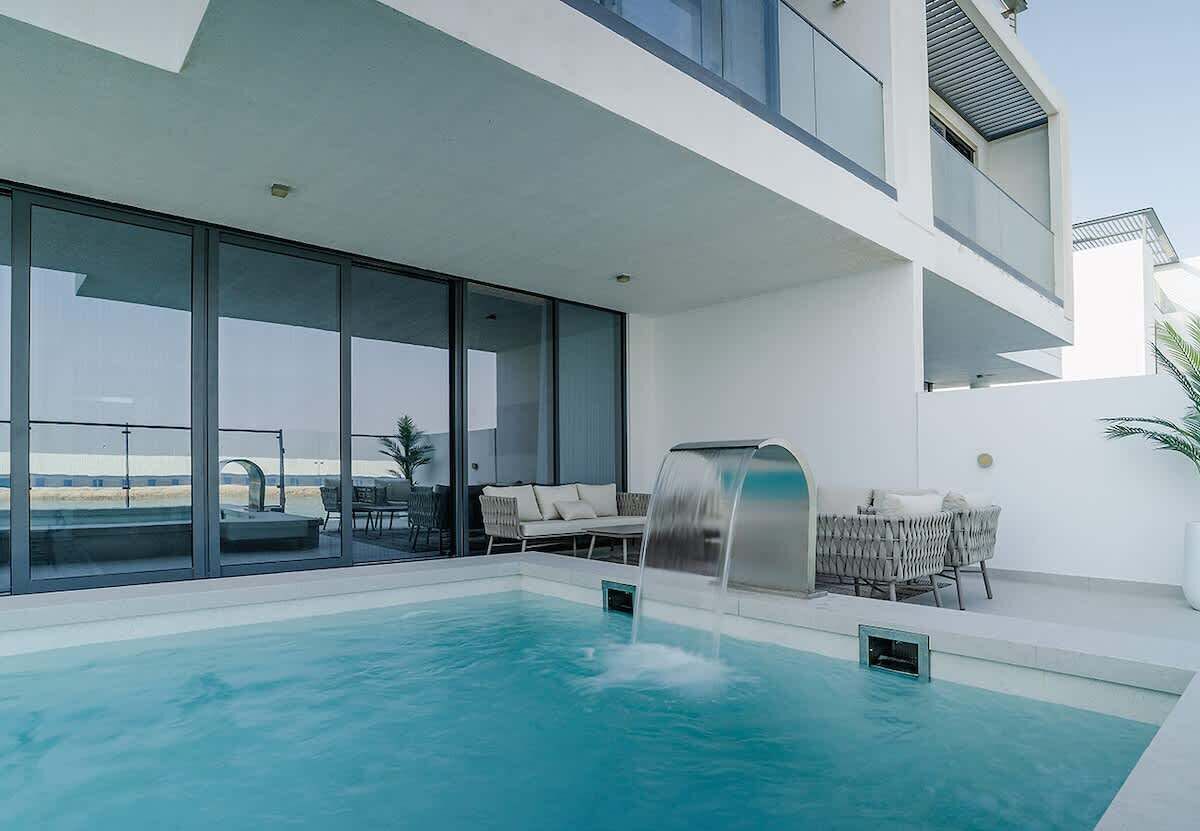 A modern pool area with a waterfall feature, a lounge chair, and a grill.