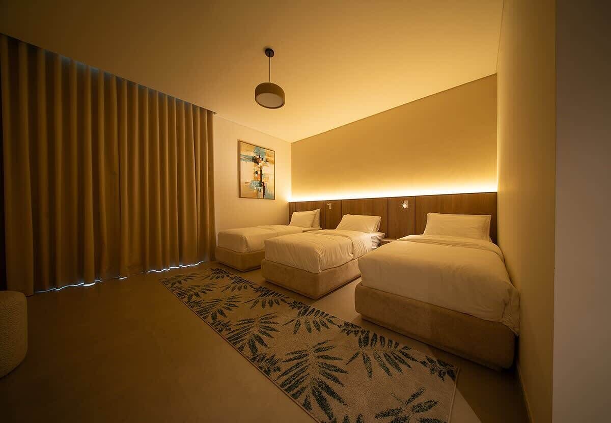 Quarto de hotel com duas camas, uma janela grande com cortinas, um quadro na parede e uma luminária no teto.