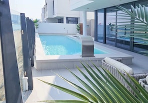 A modern outdoor pool area with a hot tub, surrounded by a white building and a glass wall.