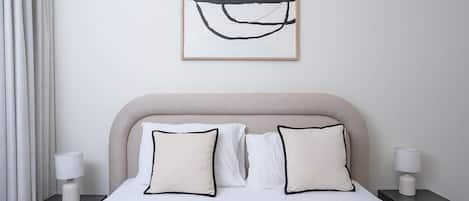 Habitación de hotel con cama, dos mesitas de noche, lámpara, cuadro abstracto enmarco y ventana con cortinas.
