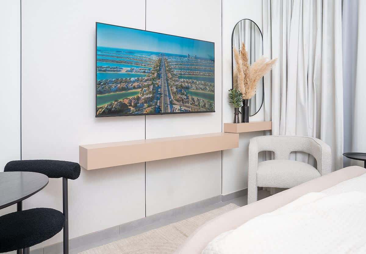 A modern hotel room with a large window, a bed, a desk, a chair, and a wall-mounted TV displaying a cityscape.