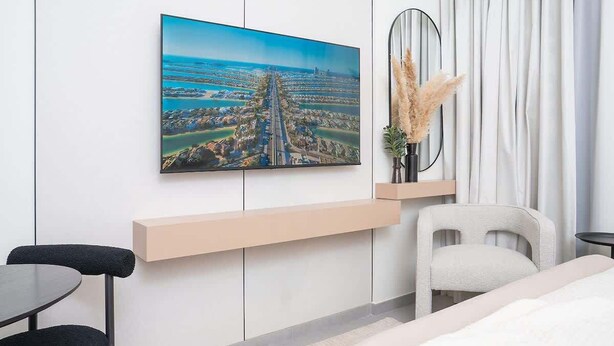 Ein modernes Hotelzimmer mit großem Fenster, einem Bett, einem Schreibtisch, einem Sessel und einem an der Wand befestigten Fernseher, der eine Stadtansicht zeigt.