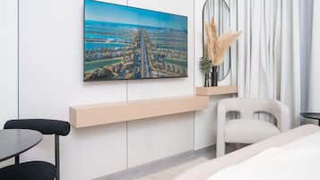 A modern hotel room with a large window, a bed, a desk, a chair, and a wall-mounted TV displaying a cityscape.