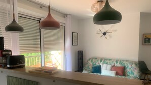 Een keuken met een houten aanrecht, een broodrooster en een koffiezetapparaat. Er is een raam met jaloezieën, een bank met bloemenprint en een wandklok.
