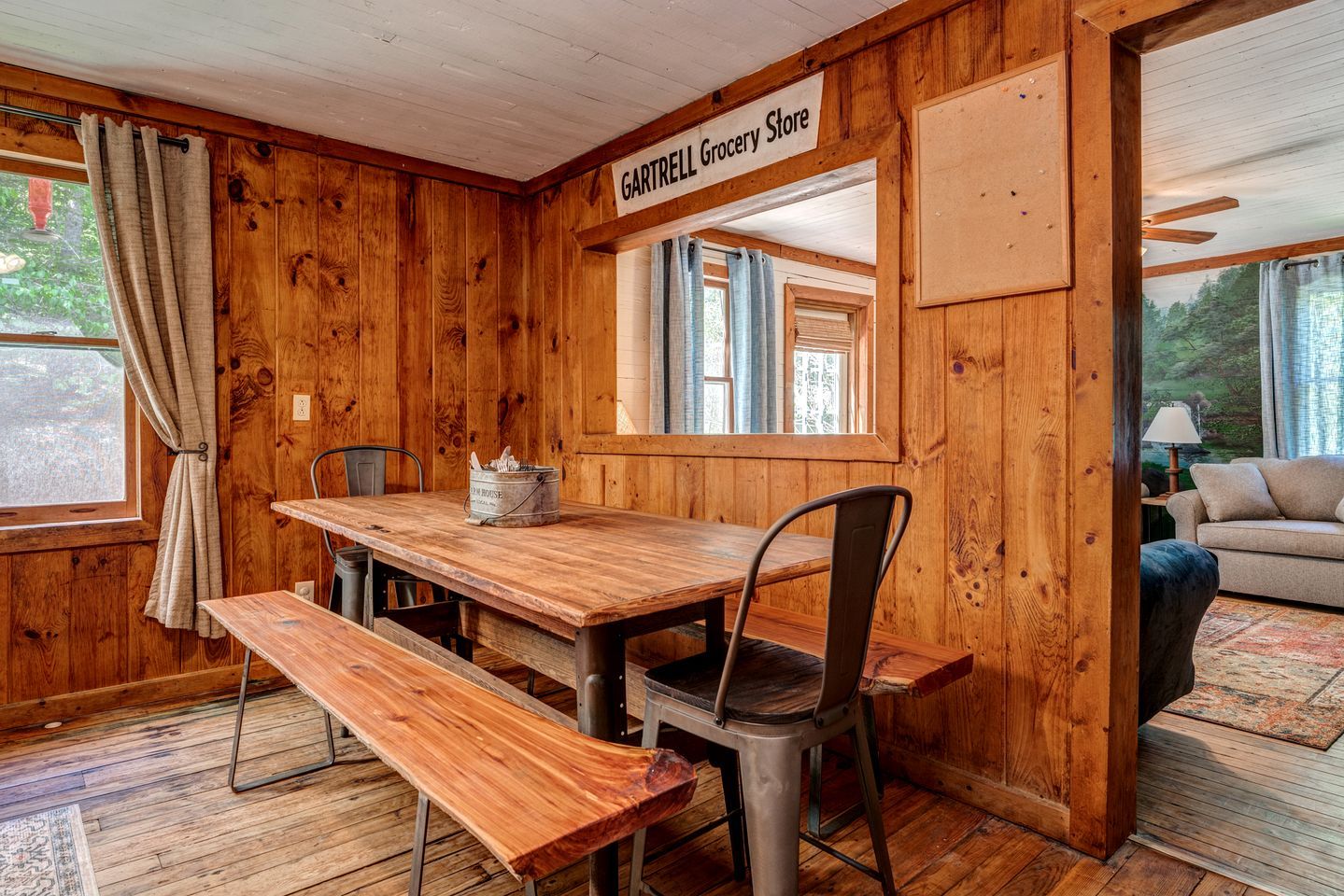 A wooden interior with a dining table, benches, a sign reading 'GARTRELL Grocery Store,' and a sofa in a room with wooden walls and windows.