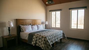 Un dormitorio con cabecera de madera, cama con colcha estampada, dos lámparas de noche y dos ventanas con persianas.