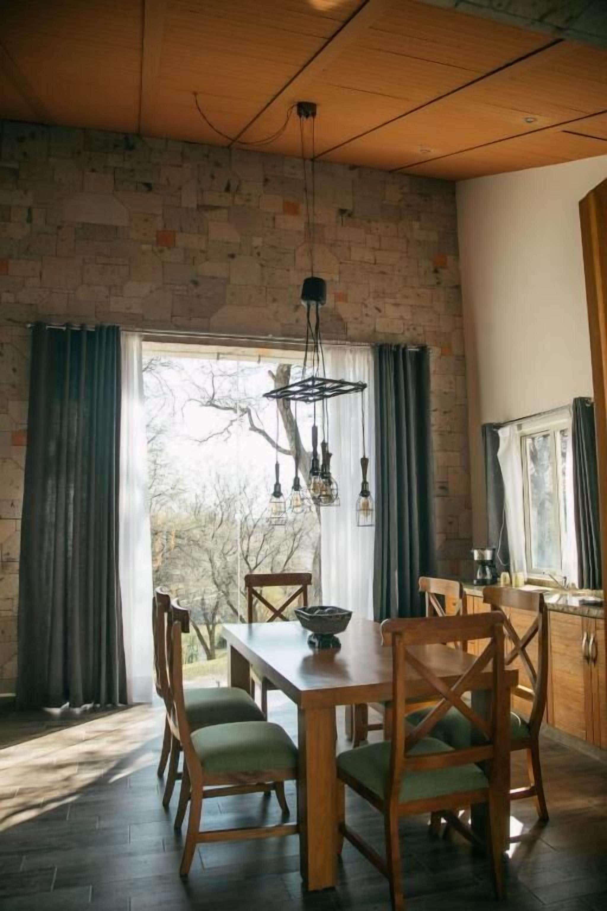 A dining area with a wooden table and chairs, a hanging light fixture, and a view through a window with curtains.
