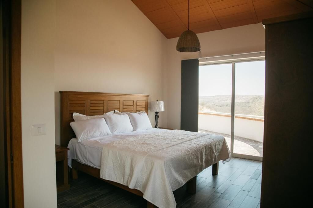 Un dormitorio con cabecera de madera, cama con ropa de cama blanca, lámpara de noche y un ventanal con vistas al paisaje.