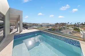 Ein moderner Pool im Freien mit freiem Blick auf die Umgebung, darunter ein Gebäude mit Balkon sowie Palmen und entfernte Strukturen.