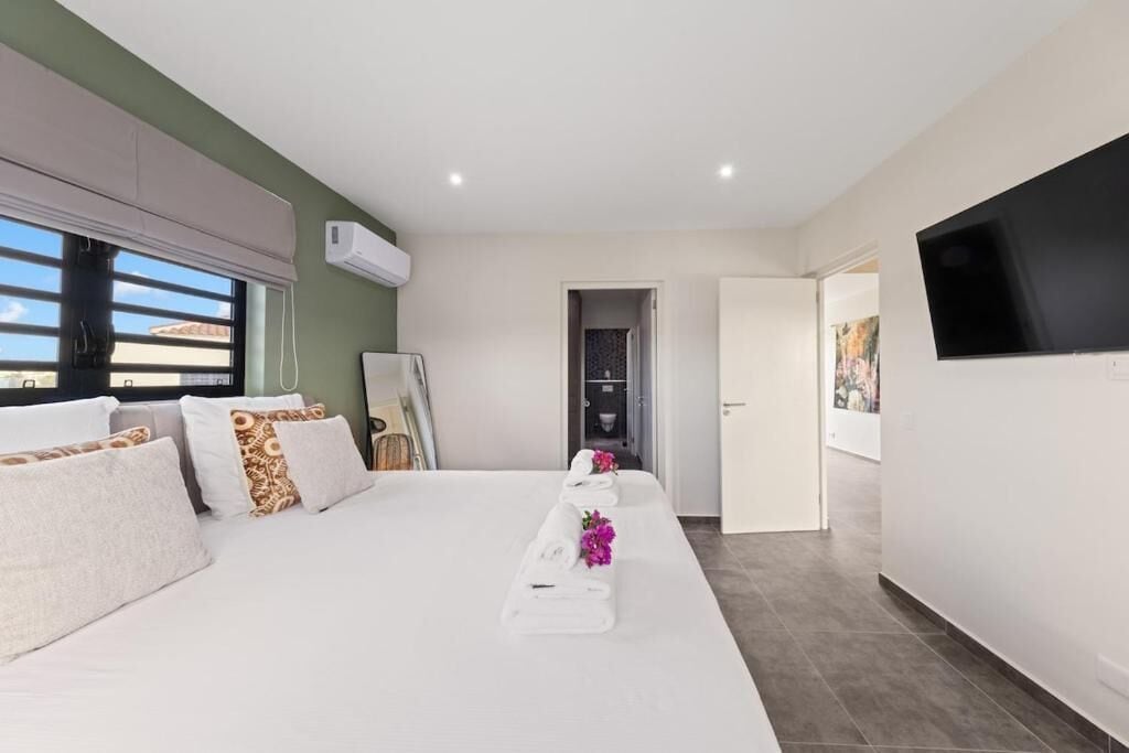 A hotel room with a bed, a flat-screen TV mounted on the wall, and a window with blinds.