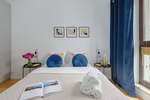 A neatly made bed with a white quilt, two blue decorative pillows, a stack of white towels, and a book on the bed. There are three framed artworks on the wall, a vase with pink flowers on a bedside table, and a blue curtain on the window. - Lumina 3-room apart near Wola Museum (Warszawa)