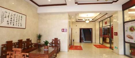 A hotel lobby with a reception desk, wooden furniture, a chandelier, and a staircase leading to the dining area.