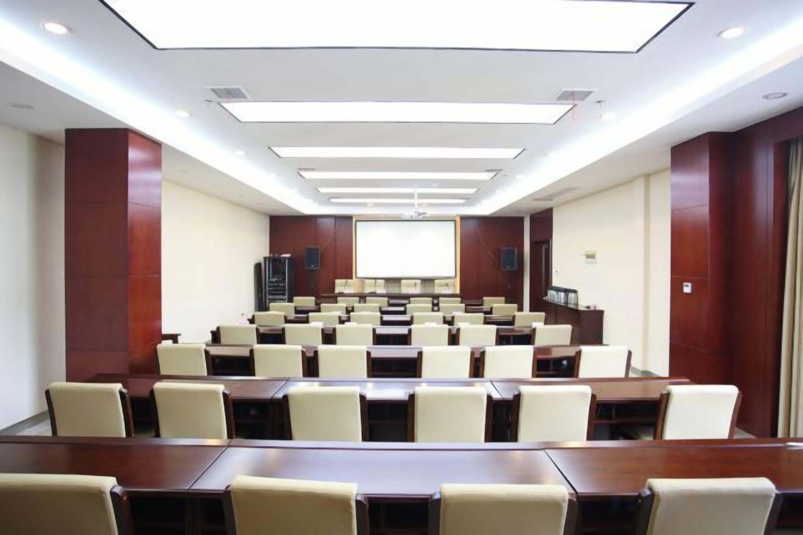 Un salón de conferencias con filas de mesas y sillas, una pantalla de proyector y una zona para presentaciones.