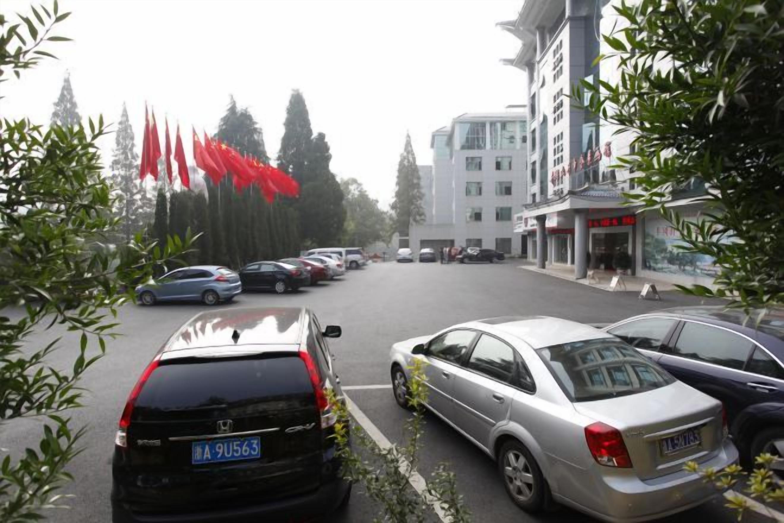 Un estacionamiento con varios coches, un edificio con un cartel y banderas rojas a la izquierda.