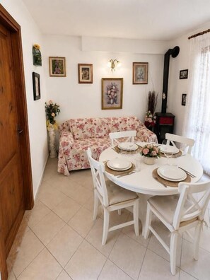 A dining area with a round table set for four, white chairs, and a floral sofa in the background. - Beach Walk Apartments - San Teodoro (San Teodoro)