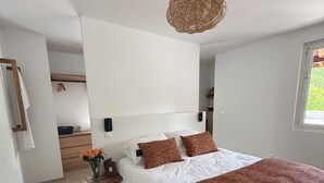 A bedroom with a large bed, a bedside table with a vase of flowers, a hanging light fixture, and a window. - SOLEO Vacation Home (Solférino)