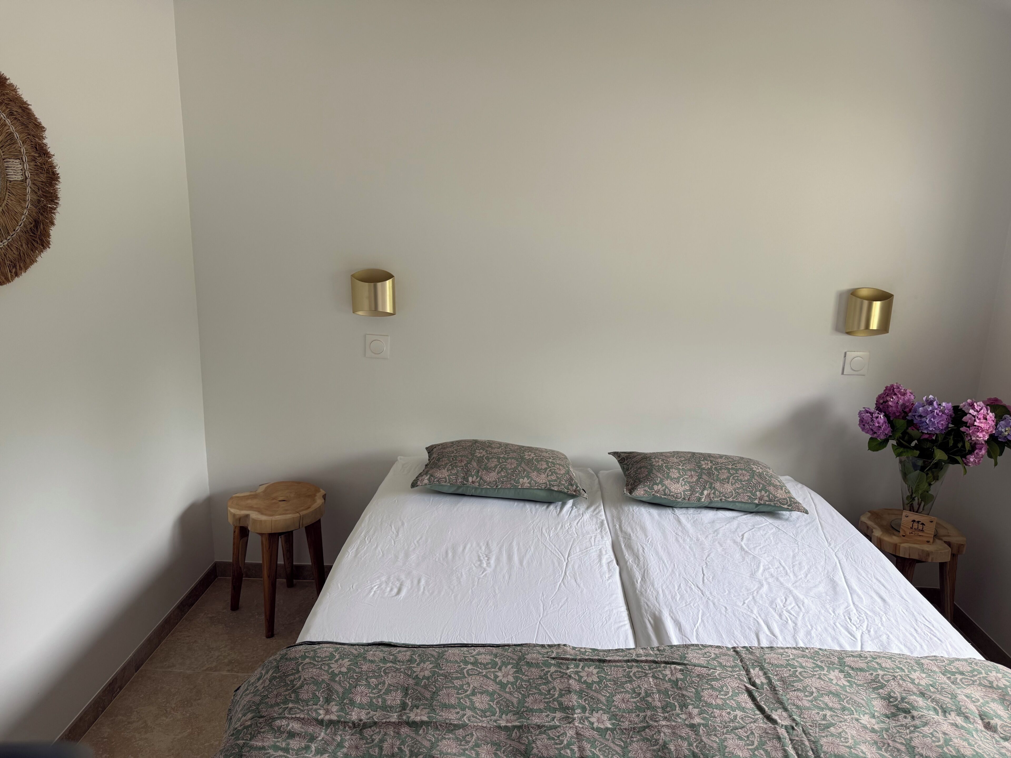 Un lit avec deux oreillers, une table de chevet ornée d’un vase de fleurs et un luminaire fixé au mur.