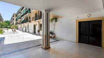 Een hotelgang met een betegelde vloer, een centrale kolom met een decoratief patroon, een deur met een zwart metalen hekwerk en een balkon met planten.