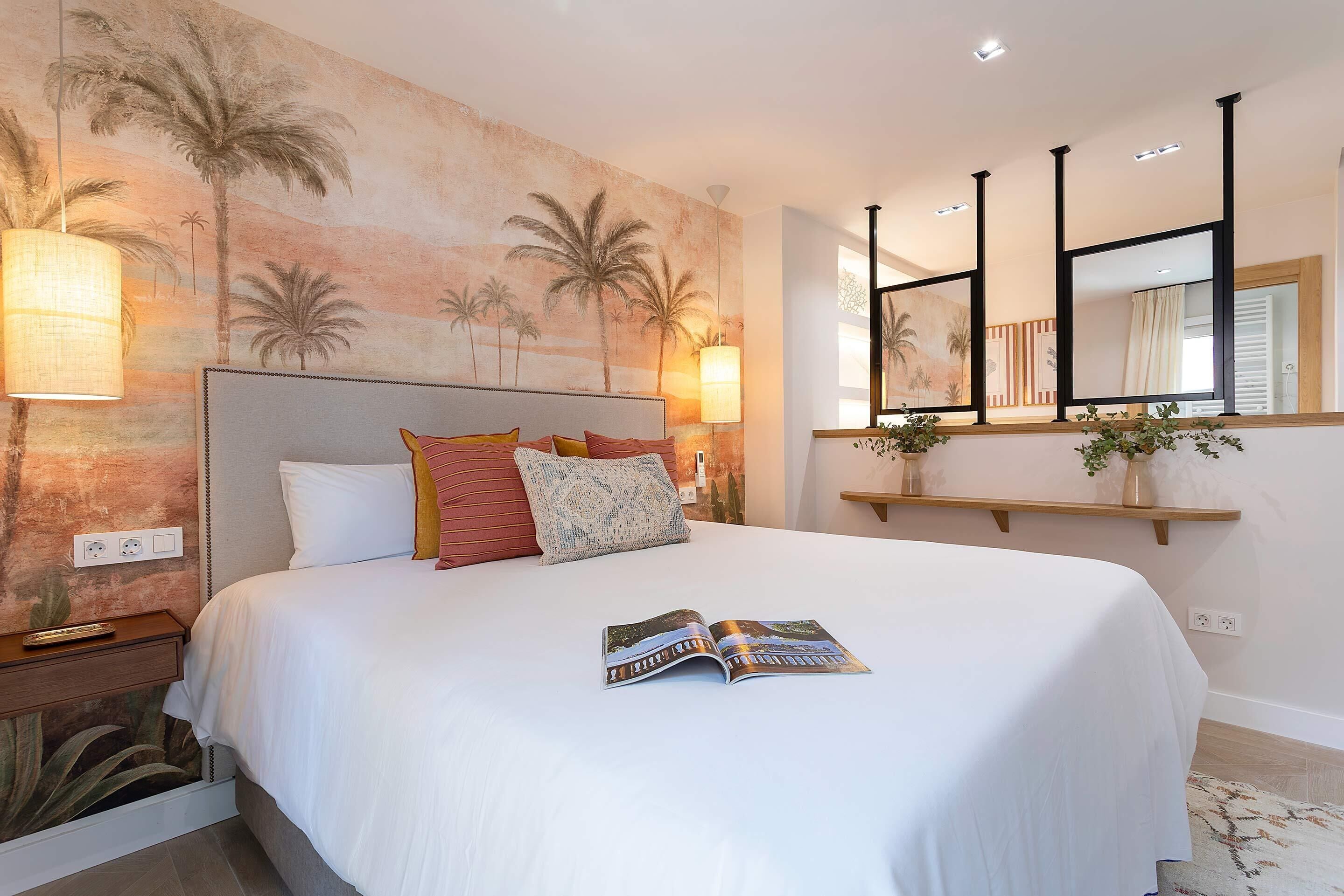 Un lit bien fait, avec un magazine à portée de main, une tête de lit ornée d’une fresque représentant un palmier, des lampes de chevet et une étagère en bois sur laquelle est posée une plante.