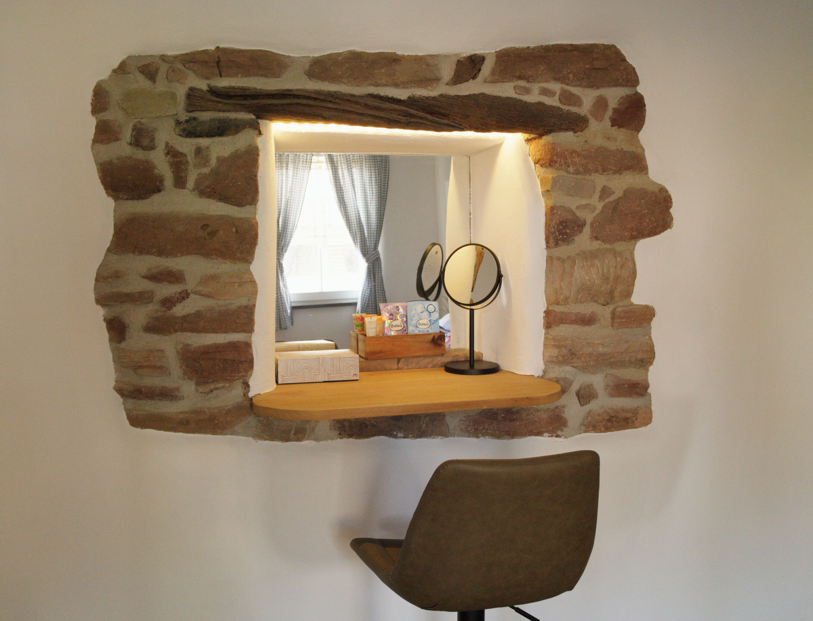 A stone-framed mirror with a light above it, a chair, and a window with curtains.