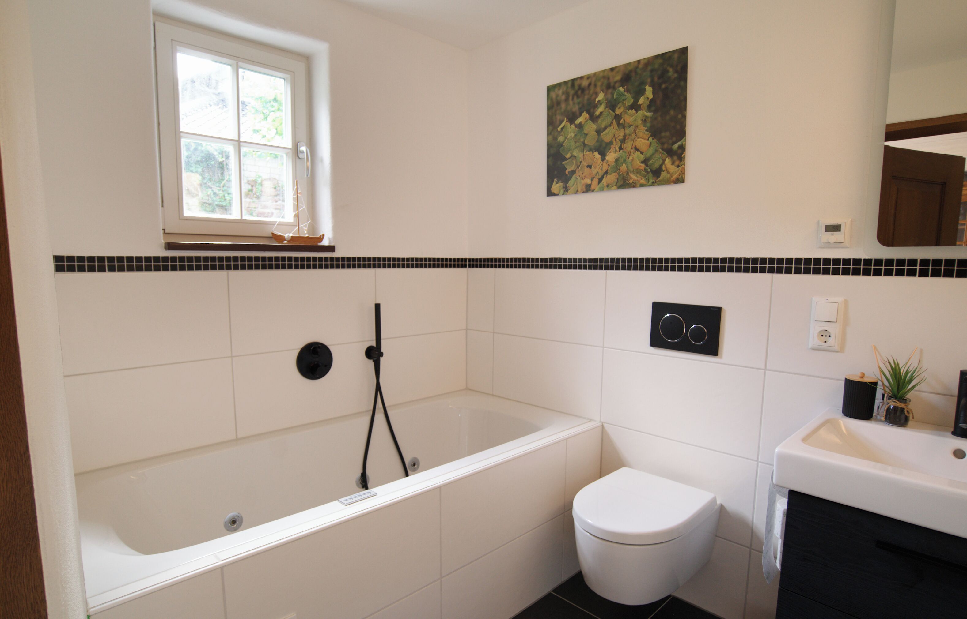 Ein Badezimmer mit Badewanne, Toilette und Waschbecken, mit einem Fenster, gefliesten Wänden und einem Bild.