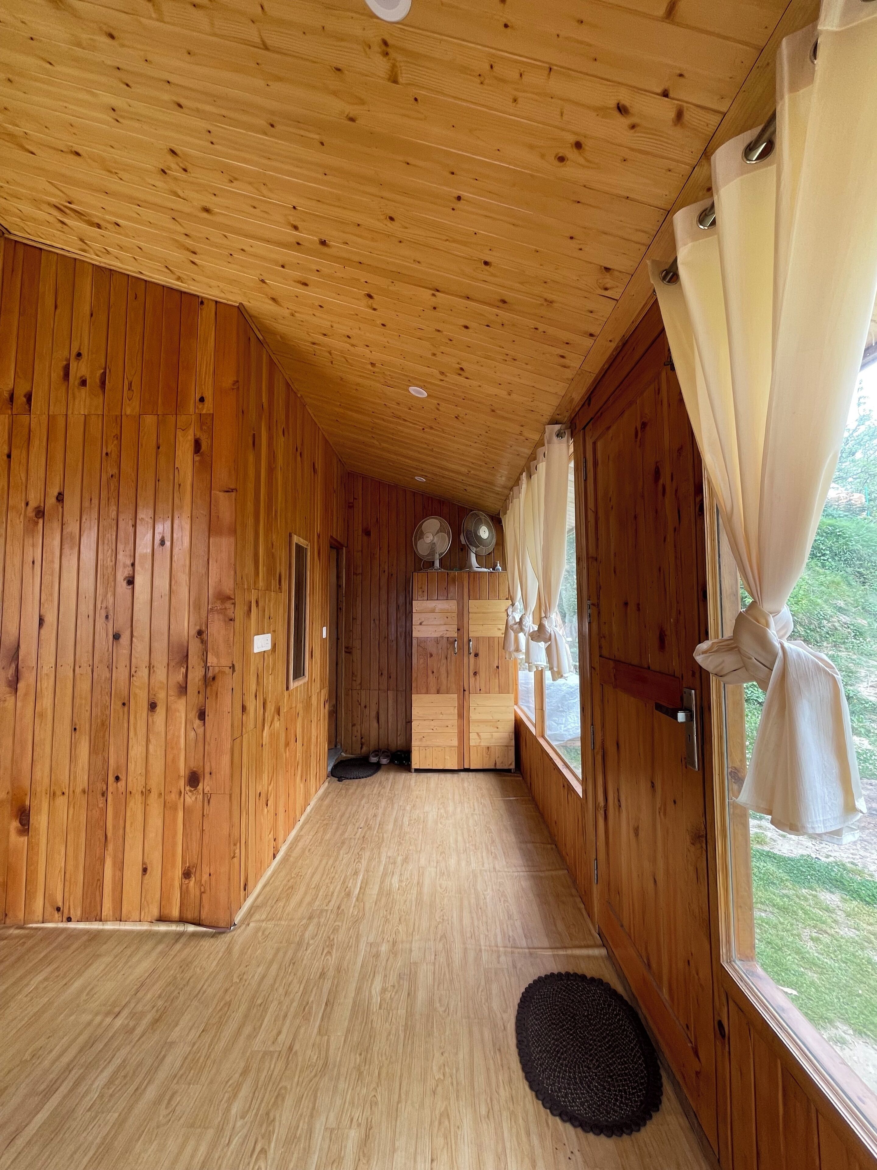 A wooden interior hallway with a door leading to a room, a window with curtains, and a pair of shoes on a mat.