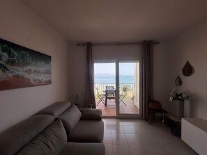 Ein Wohnzimmer mit einer Couch, einem Gemälde, einem Balkon mit Tisch und Stühlen und Blick aufs Meer.