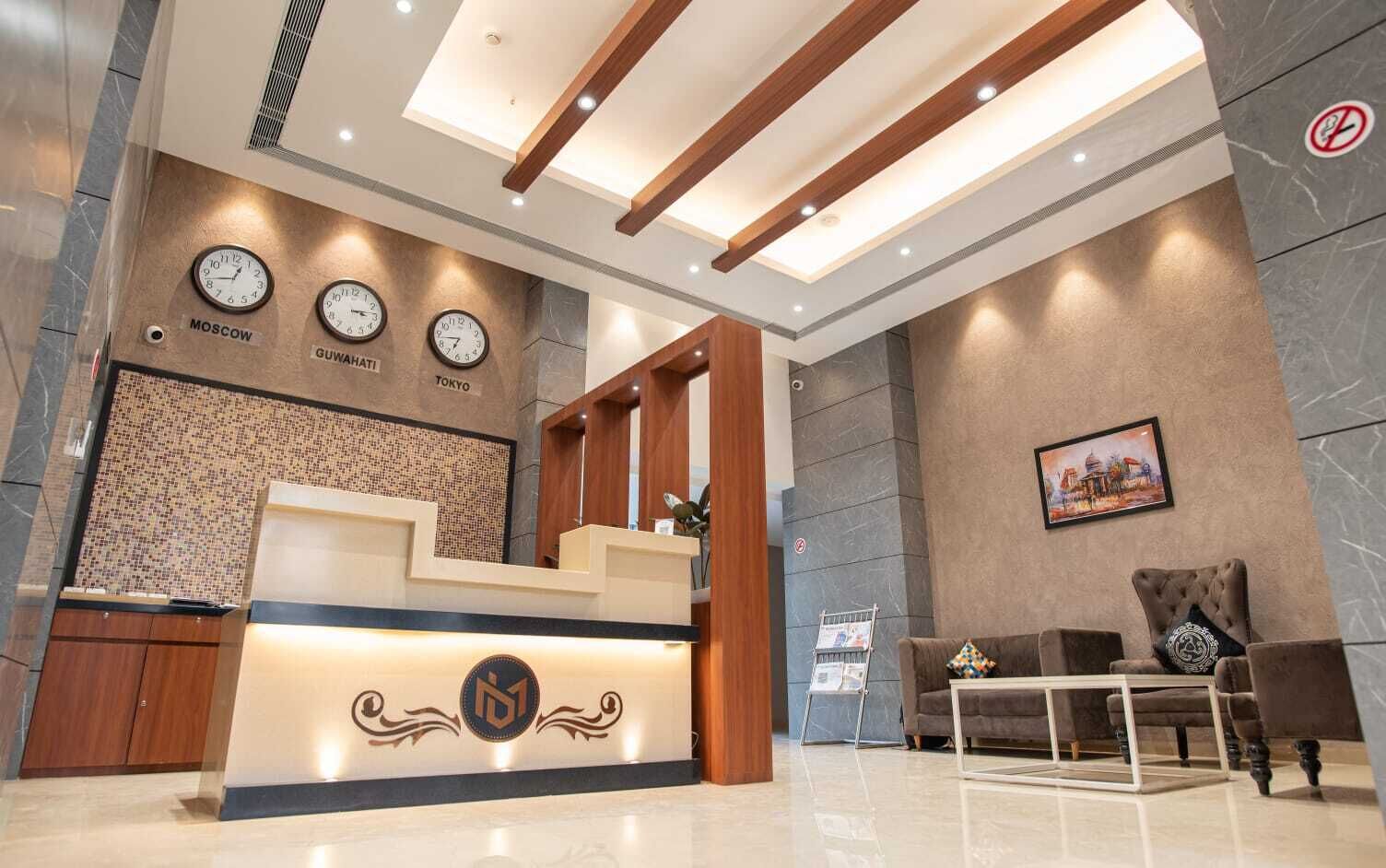 Zona de recepción de un hotel con mostrador, relojes mostrando la hora en Moscú, Guwahati y Tokio, un área de descanso con sofá y sillones, y una pared con un cuadro enmarcado.
