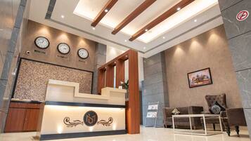 Zona de recepción de un hotel con mostrador, relojes mostrando la hora en Moscú, Guwahati y Tokio, un área de descanso con sofá y sillones, y una pared con un cuadro enmarcado.
