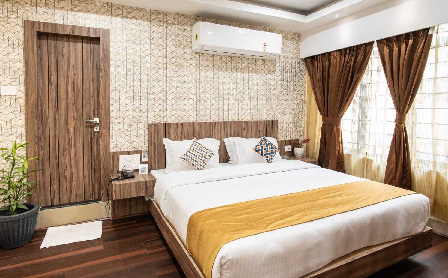 Habitación de hotel con cama, mesita de noche, cortinas y una planta en maceta, además de un equipo de aire acondicionado.