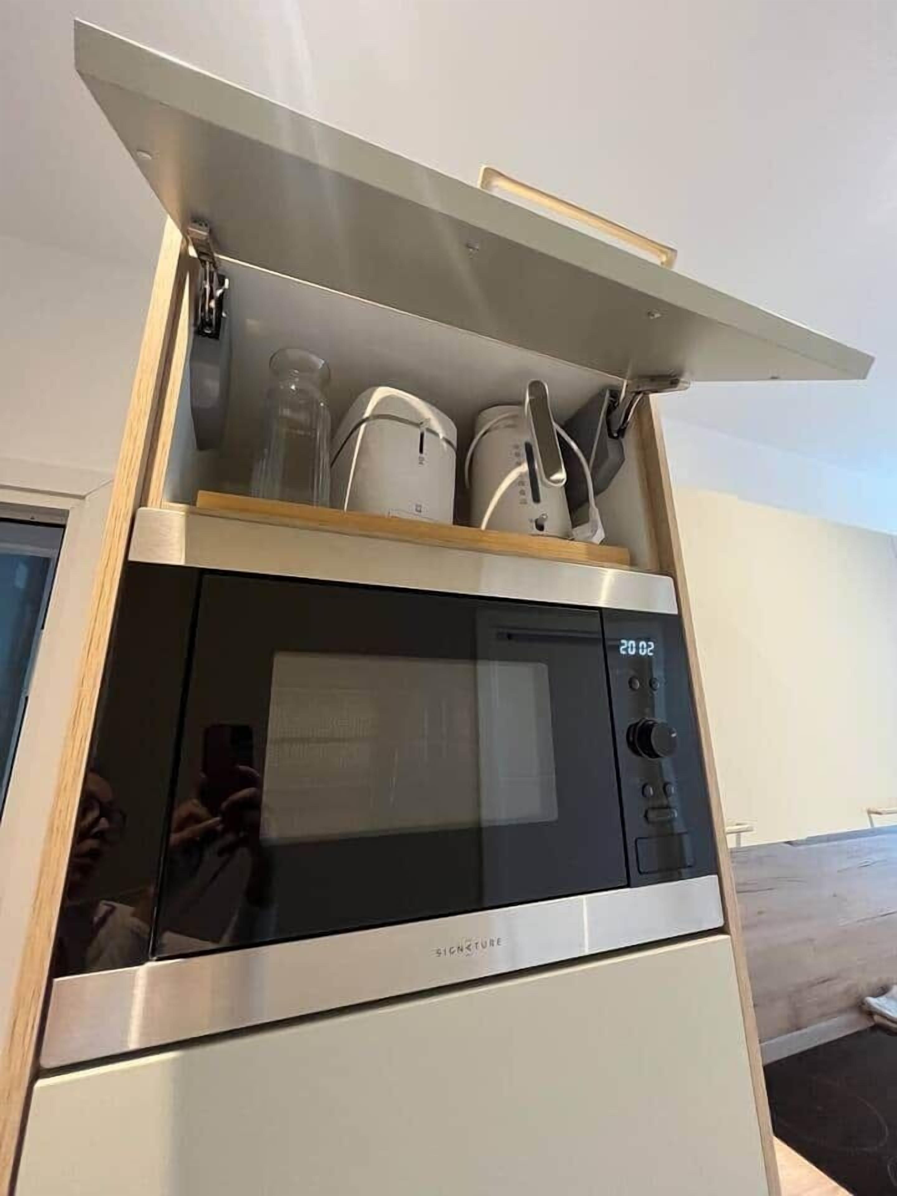 Een keuken met een magnetron, oven en een houten plank met keukengerei.