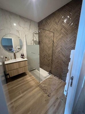 Un baño moderno con pared de diseño en espiga, suelo de madera, cabina de ducha de vidrio y lavamanos blanco con espejo.
