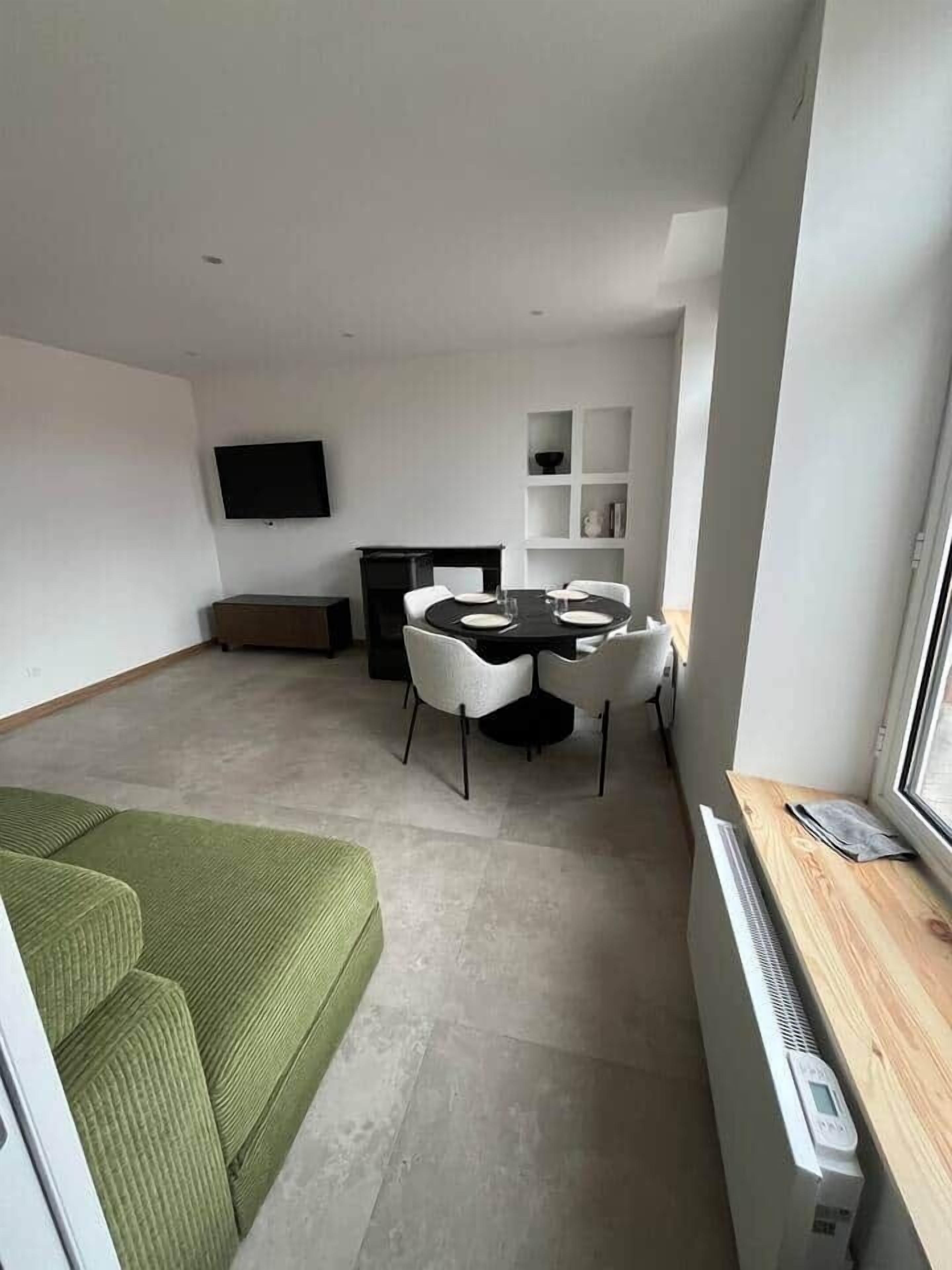 Un espace de vie compact comprenant un coin repas, un téléviseur à écran plat fixé au mur, un canapé vert et une fenêtre donnant sur un paysage agréable.
