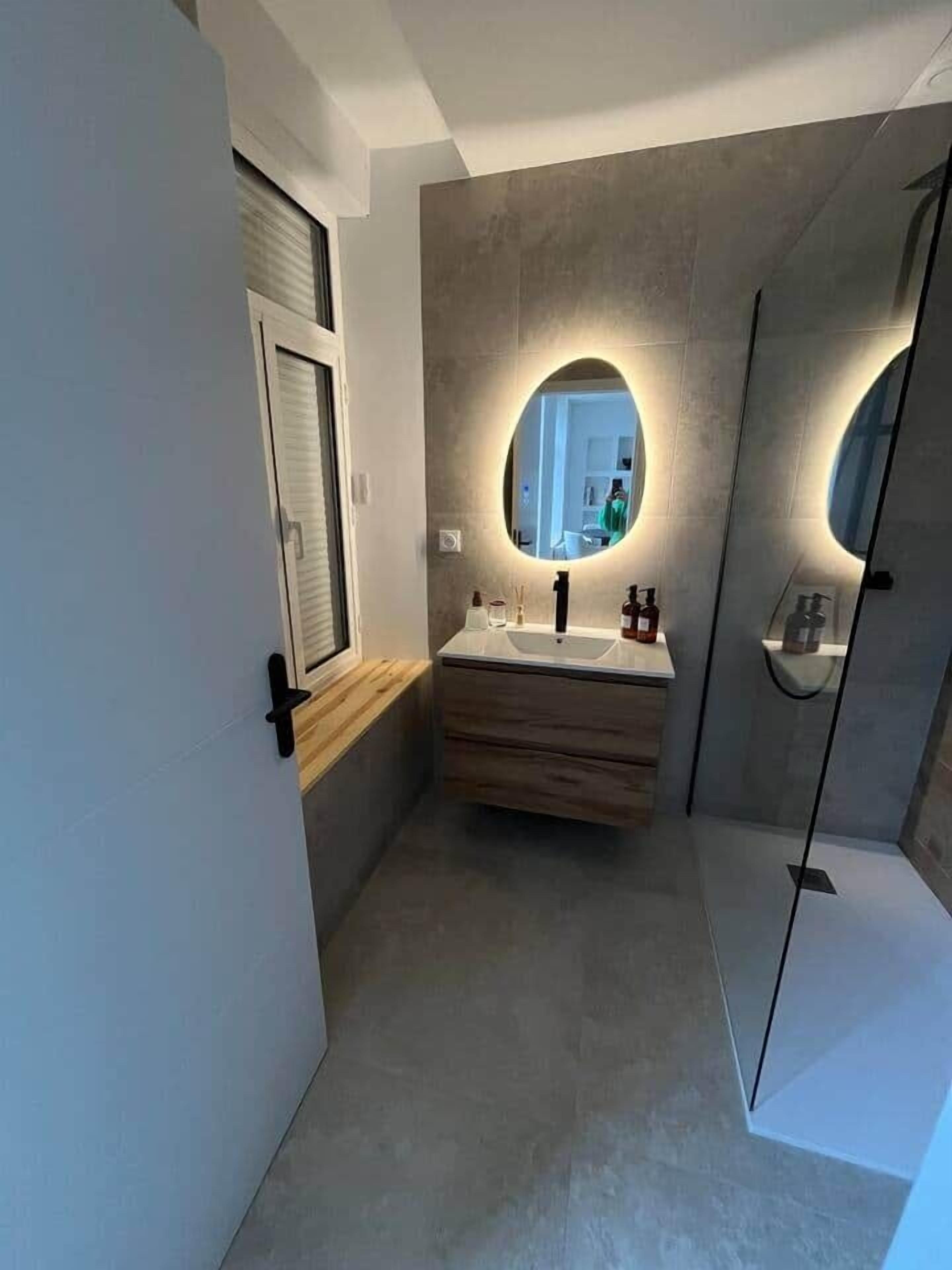 Une salle de bain moderne avec un meuble-lavabo en bois, un grand miroir rond doté d’un éclairage indirect, une cabine de douche en verre et une fenêtre bien visible.