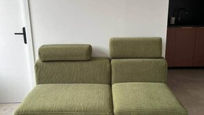 Un canapé modulable avec un coussin et un pouf assorti, dans une pièce où l’on aperçoit une porte blanche et, au fond, un coin cuisine.