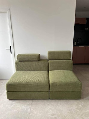 A sectional sofa with a cushion and a matching ottoman in a room with a white door and a kitchen area in the background. - Bayt al Nour | Designer & Cozy Apartment (Givet)