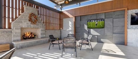 Une terrasse extérieure dotée d’une cheminée en pierre, de meubles en osier et d’un écran de télévision diffusant un événement sportif.