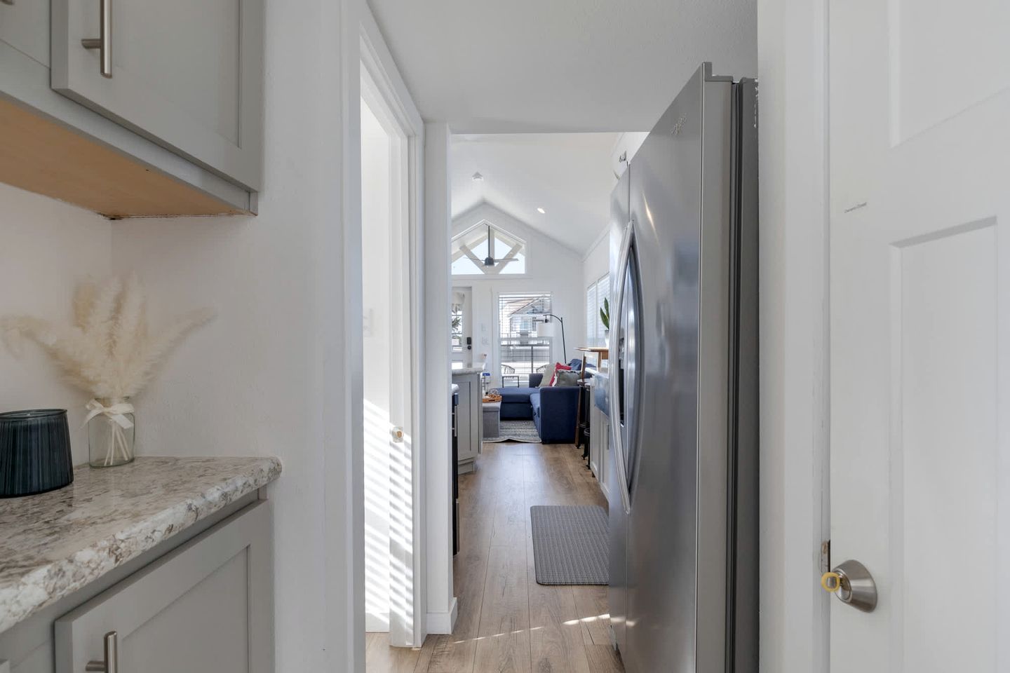A narrow hallway with a kitchen area on one side, a dining area with a blue sofa, and a view into a room with a staircase.