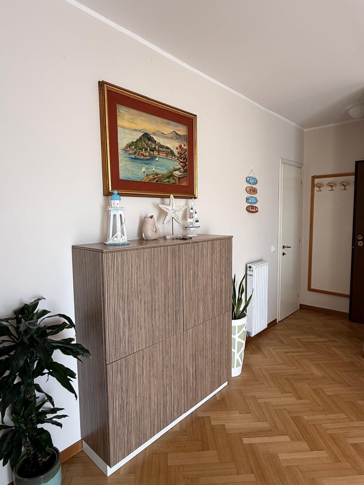 A room with a wooden cabinet, a painting of a coastal scene, a potted plant, and a radiator.