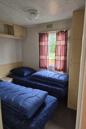Ein kleiner, kompakter Raum mit zwei Betten, einem Fenster mit Vorhängen, einer Deckenleuchte und einem Kleiderschrank.