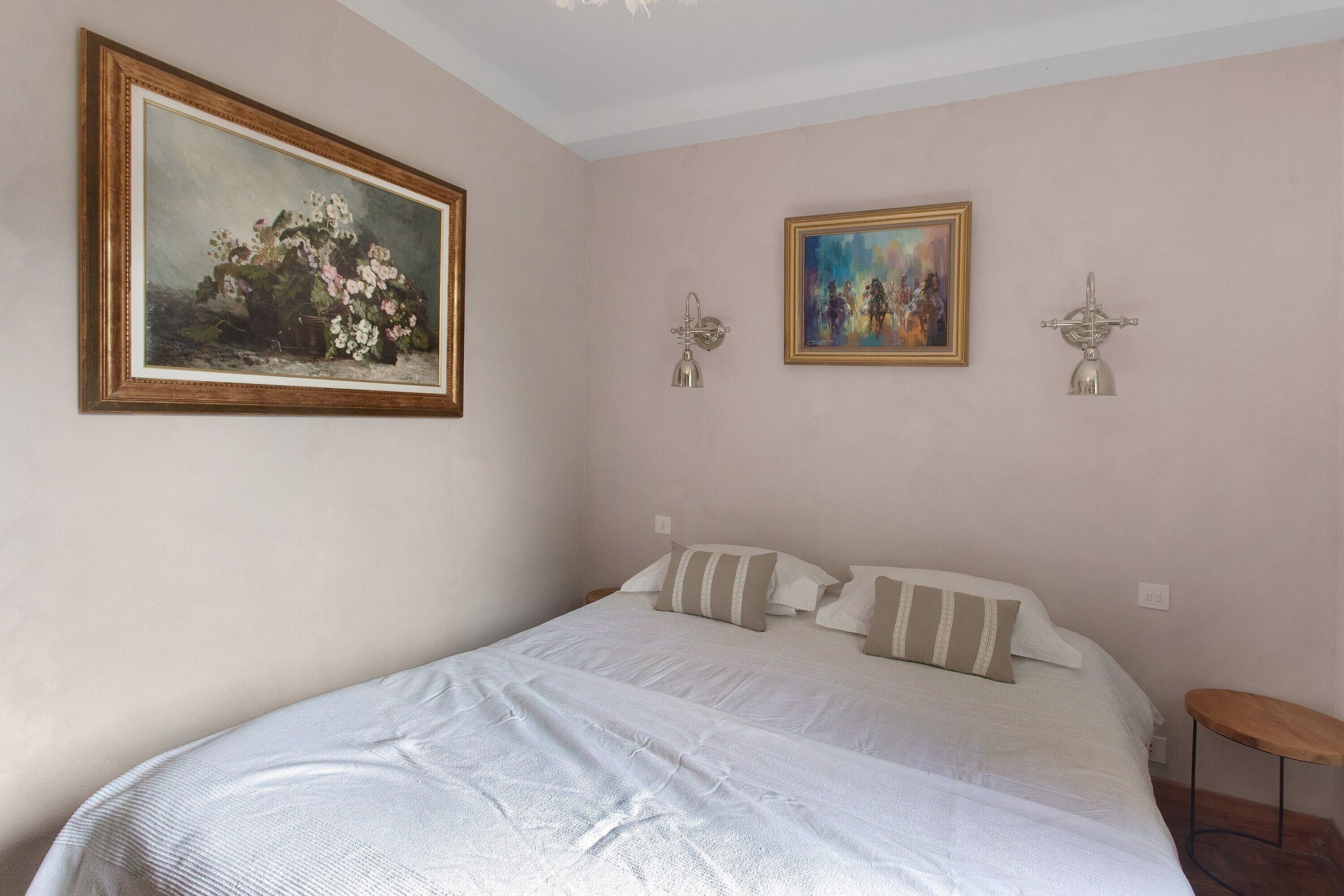 A bedroom with a large bed, two wall-mounted lamps, and two framed artworks.
