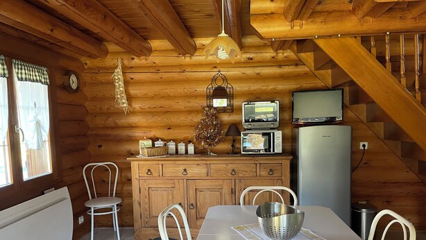 A wooden cabin interior with a dining table, chairs, a kitchen area with appliances, and a staircase.