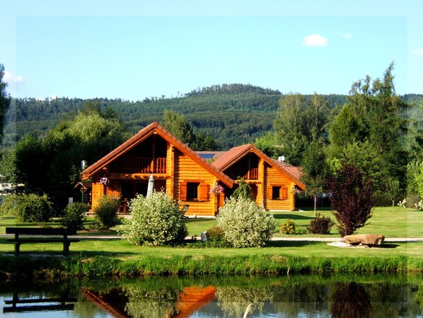 A wooden cabin with a balcony, surrounded by a pond, trees, and a grassy area.