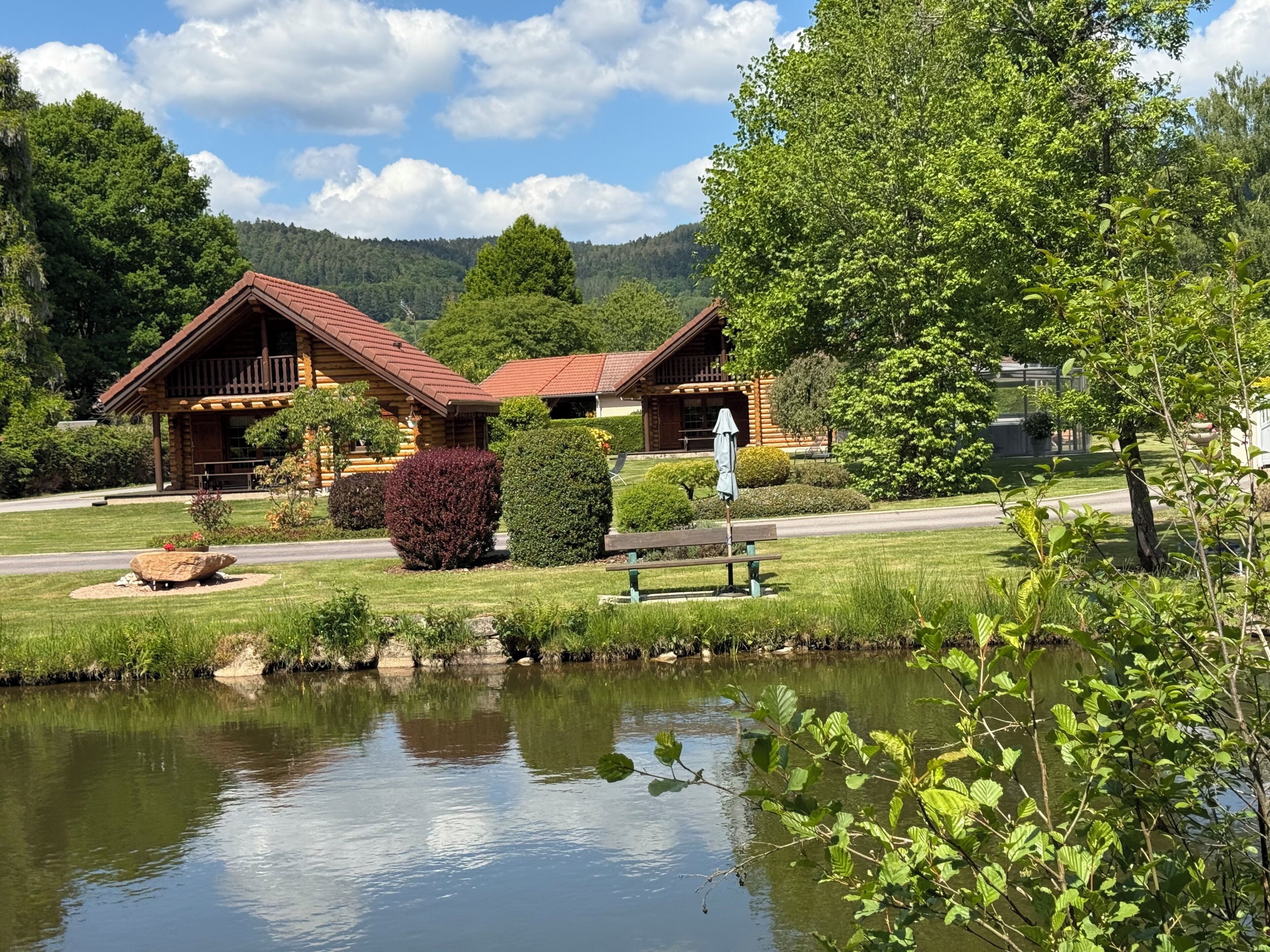 A log cabin hotel with a pond in front, surrounded by greenery and hills.
